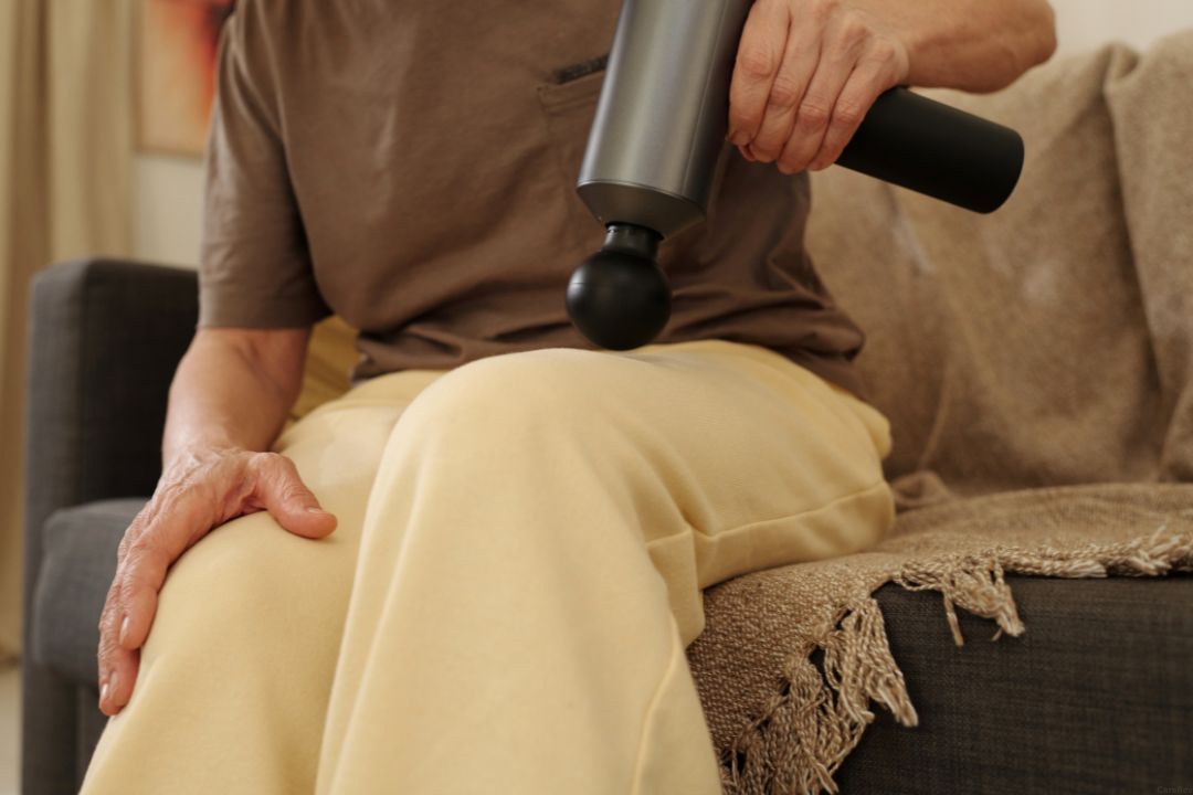 An older person uses a massage gun on their thigh muscle while sitting on a couch.