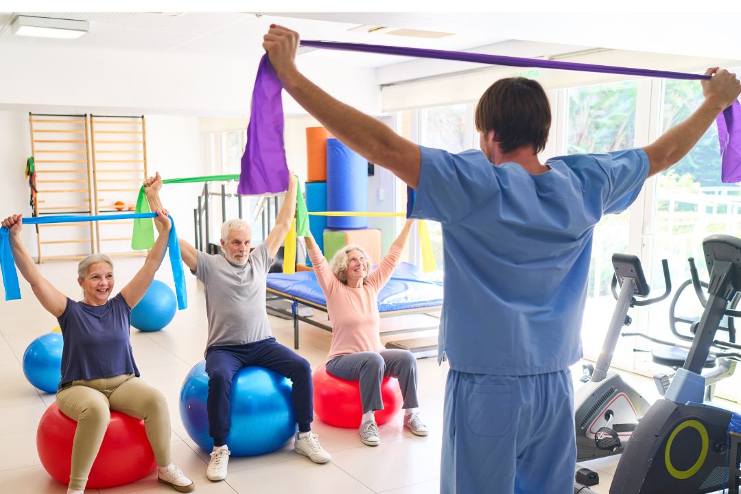 A physical therapist holds a resistance band over his head in front of three patients sitting on stability balls.