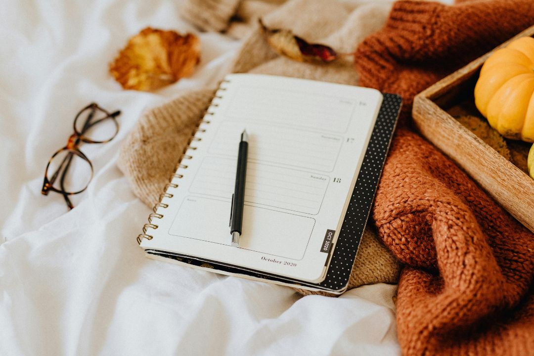 A daily planner sits on top of blankets with a pen and glasses near by.