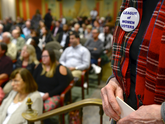 A close up of a League of Women Voters pin on a woman in a plaid scarf, with a blurred audience in the background