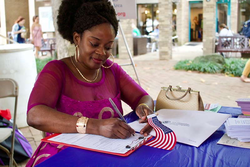 A black woman in a pink dress sitting at a table filling out a voter registration form with a small American flag in her hand