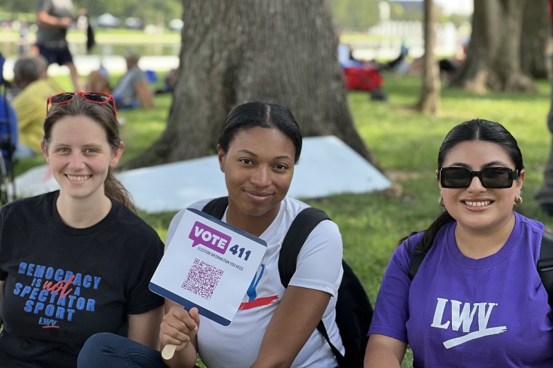three young women sitting outdoors and one is holding a Vote411 sign