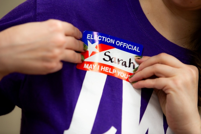 closeup of a person in a purple LWV shirt putting on a red white and blue name badge that says "election official"