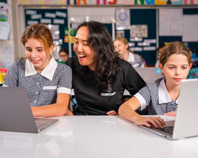 A Code Camp educator with two primary students coding on laptops in class