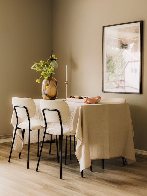 Spisebord med beige duk og svarte stoler med hvitt trekk, dekket på med en glassvase i svart og gult dekorativt mønster med en syregrønn kvist, med kiwi på et fat. Bildet over bordet på veggen.