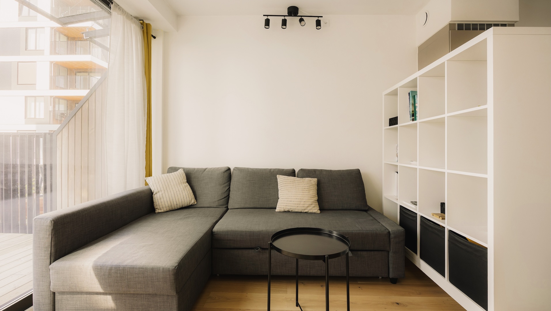 Leilighet med lyse vegger og sorte spotlights i taket. Gråsort sofa med chaiselong. To lyse puter i sofaen. Lite rundt sort sofabord. Hvit hylle som romdeler på venstre side.