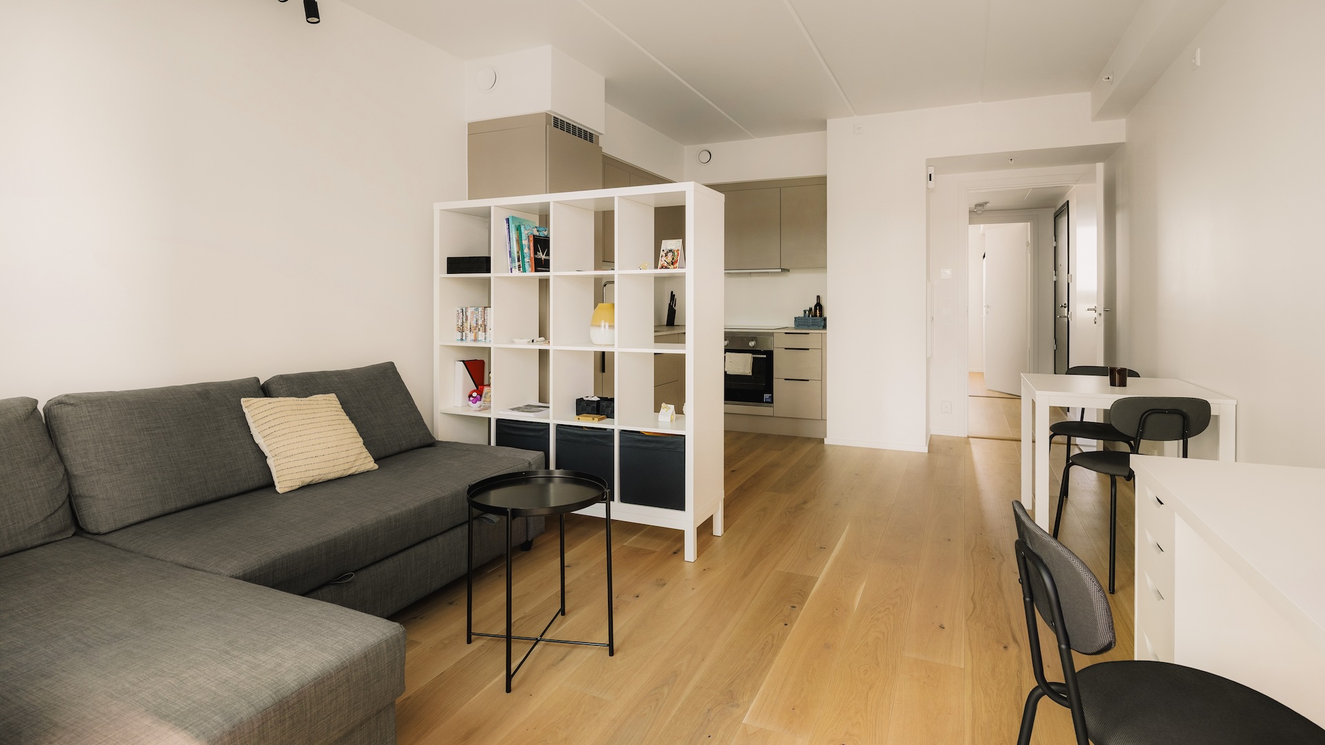 Leiligheten sett fra balkongen. Gråsort sofa på venstre side med hvit hylle som romdeler mot kjøkkenet. Hvitt spisebord med to sorte spisestoler på høyre side.