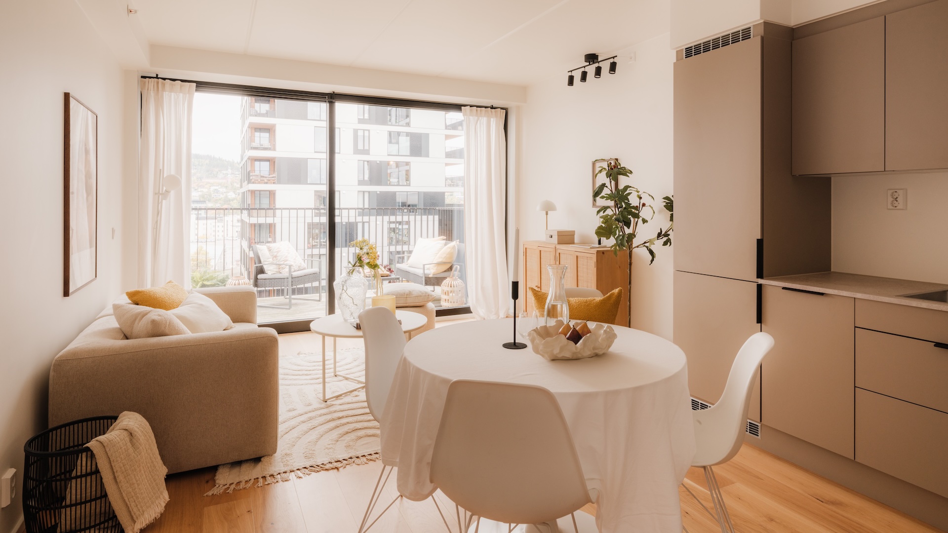 Leilighet med åpen løsning fra kjøkken til stue. Rommet er innredet med beige kjøkkeninnredning til høyre, rudt lite spisebord med spisestoler, dekor på bordet med vase, lysestake g fat. I bakgrunnet på venstre side er det innredet med sofa i lys tekstil. Veggbilde henger over sofaen- store vindusflater ut mot balkong i bakgrunnen .