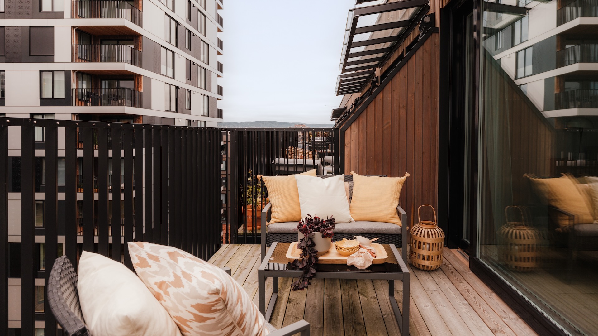 Bilde av innredet balkong tilknyttet leilighet med imprignert trgulv og sort metallrekverk. Utsikt mot høyblokker og blå himmel. Balkongen er innredet med en liten to seters sofa og lenestol i sort metall. Lyse tekstiler i puter i gult og offwhite. Plante og lykt på gulv ved siden av stol. Bord med dekor foran sofa.