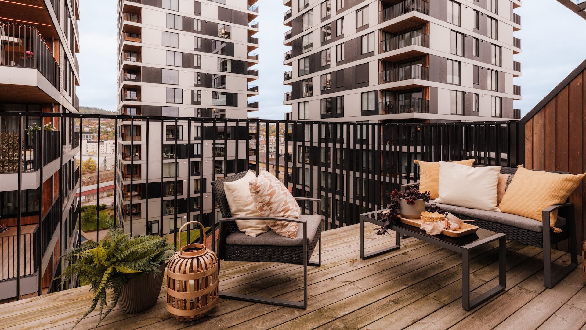 Bilde av innredet balkong tilknyttet leilighet med imprignert trgulv og sort metallrekverk. Utsikt mot høyblokker og blå himmel. Balkongen er innredet med en liten to seters sofa og lenestol i sort metall. Lyse tekstiler i puter i gult og offwhite. Plante og lykt på gulv ved siden av stol. Bord med dekor foran sofa.