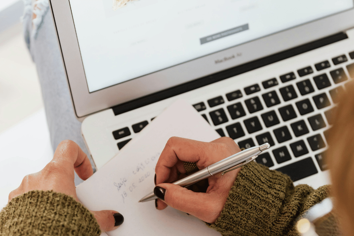 Person taking notes next to a laptop with website on screen.