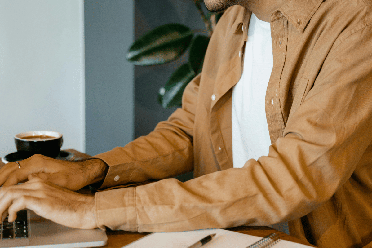 Man in a tan jacket typing on a laptop with a coffee cup nearby.