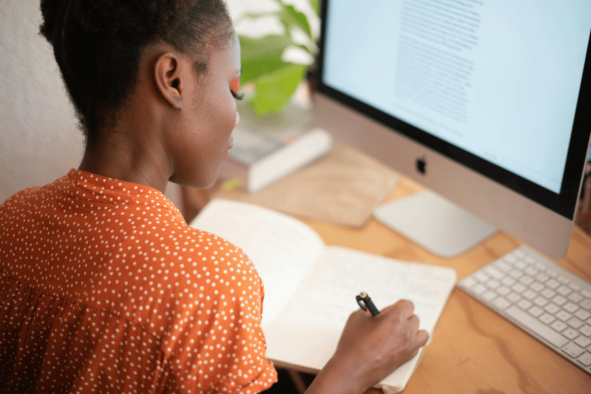 Young woman in orange polka-dot shirt writing notes from an iMac.