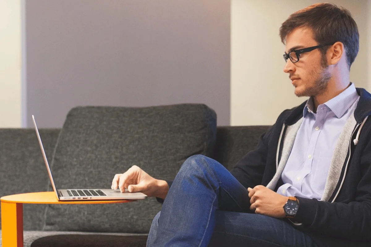 Man sitting on a couch using a laptop placed on an orange side table.