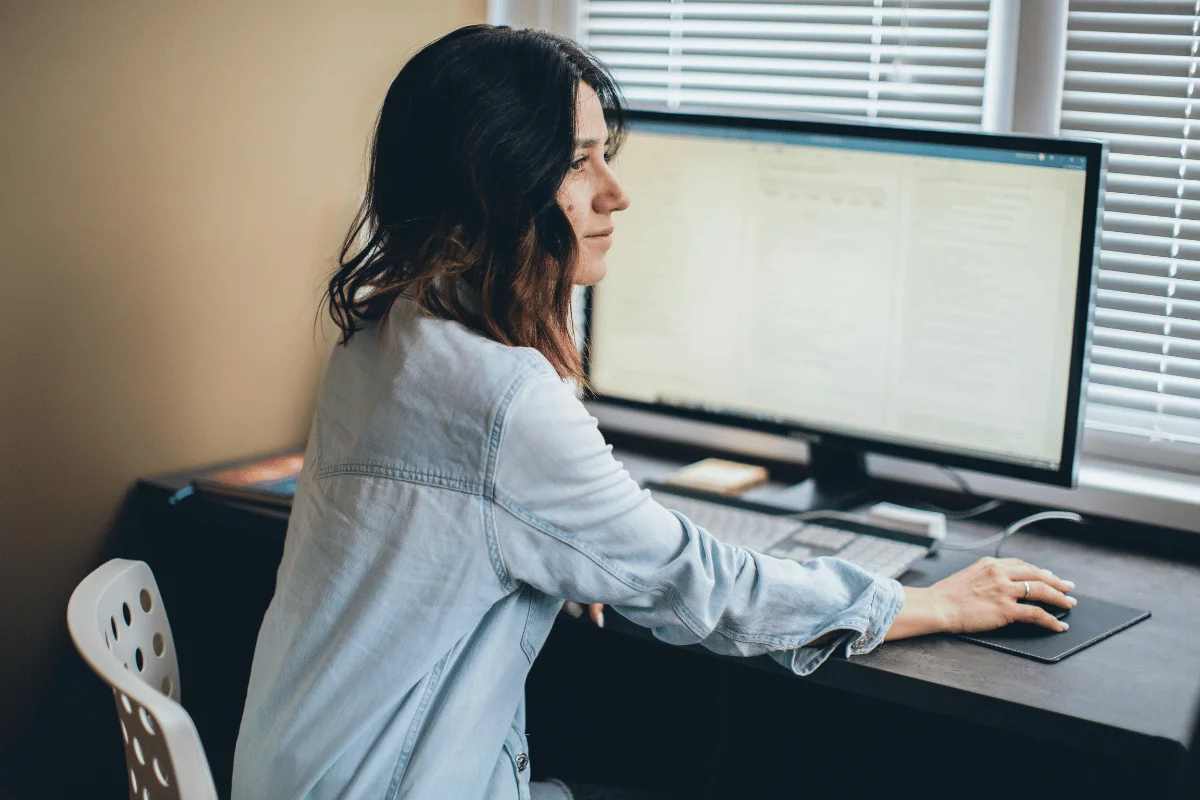 Woman working on a large desktop computer in a bright home office, focusing on her screen.