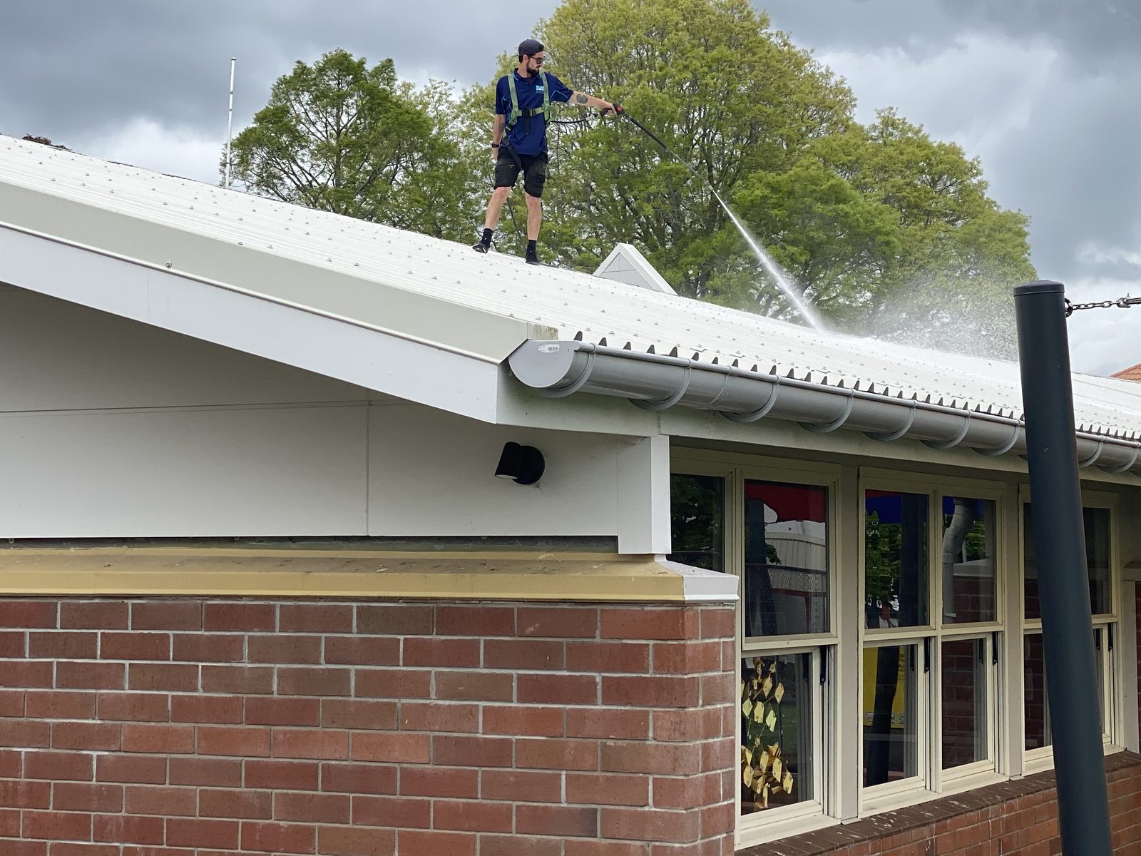 Blast Away Guys technician on a school roof in Whangarei, with colourful shade sails and a school courtyard with raised garden beds visible below