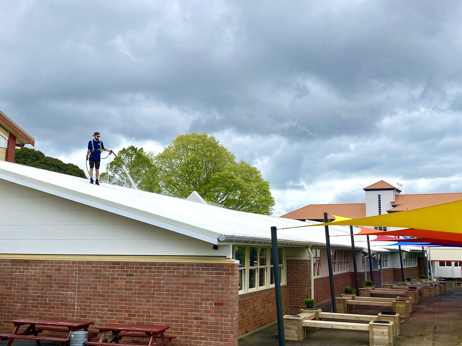 Blast Away Guys technician treating a school roof in Whangarei, standing on a white iron roof above a brick classroom building