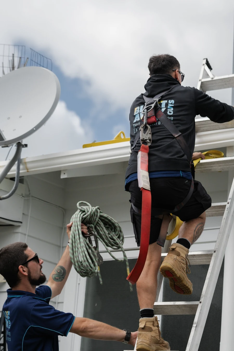 Two Blast Away Guys technicians coordinating ladder access, with one climbing and the other at the base holding a rope.