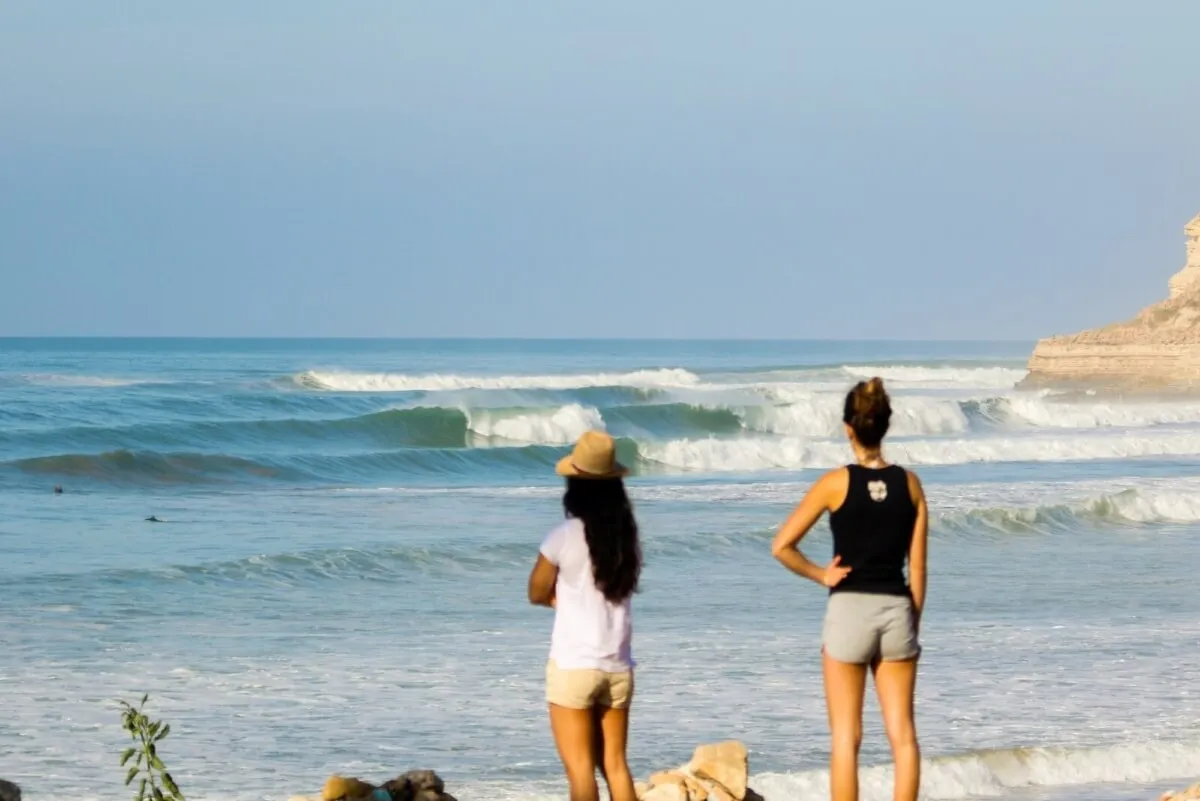 Two women standing on rocks looking at the ocean waves under a clear sky.