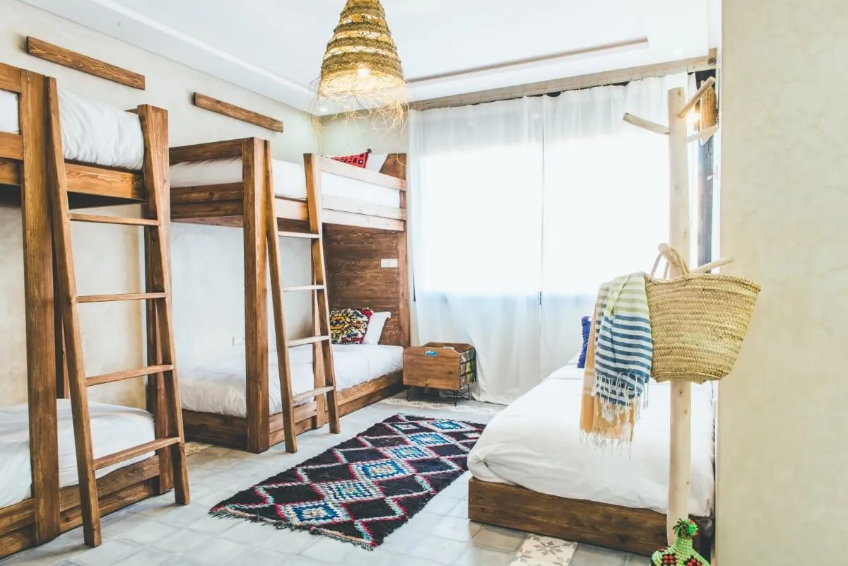 Bright room with wooden bunk beds, a single bed, patterned rug, and a woven hanging lamp.