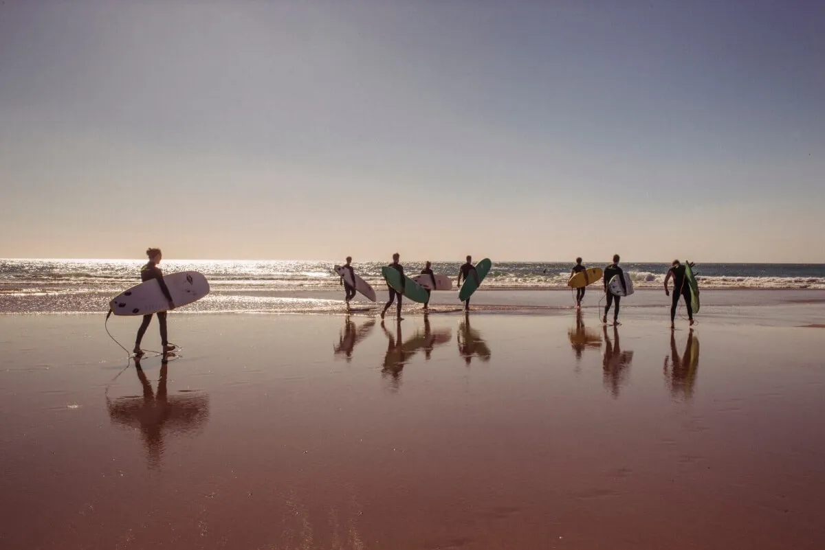 Group of surfers carrying surfboards walking towards the ocean on a reflective wet beach at sunset.