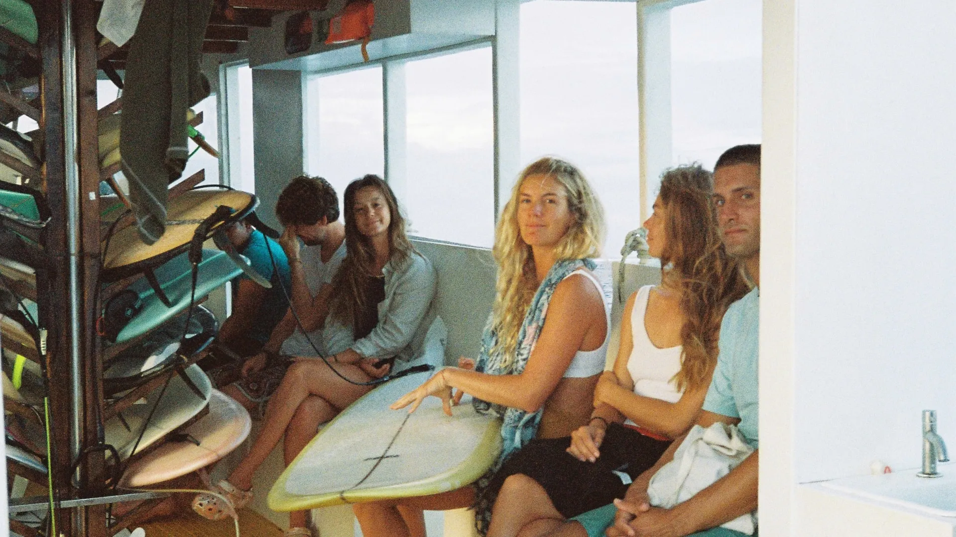 Five young adults sitting indoors near surfboards, one holding a surfboard with a towel draped over their shoulders.