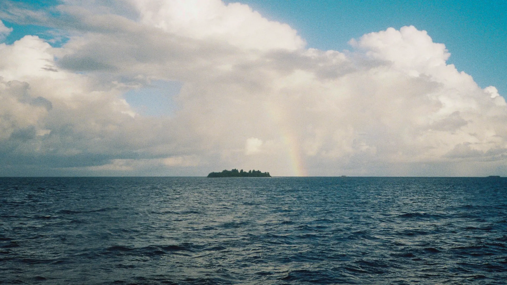 Small island surrounded by ocean water with a faint rainbow and large clouds in the sky.