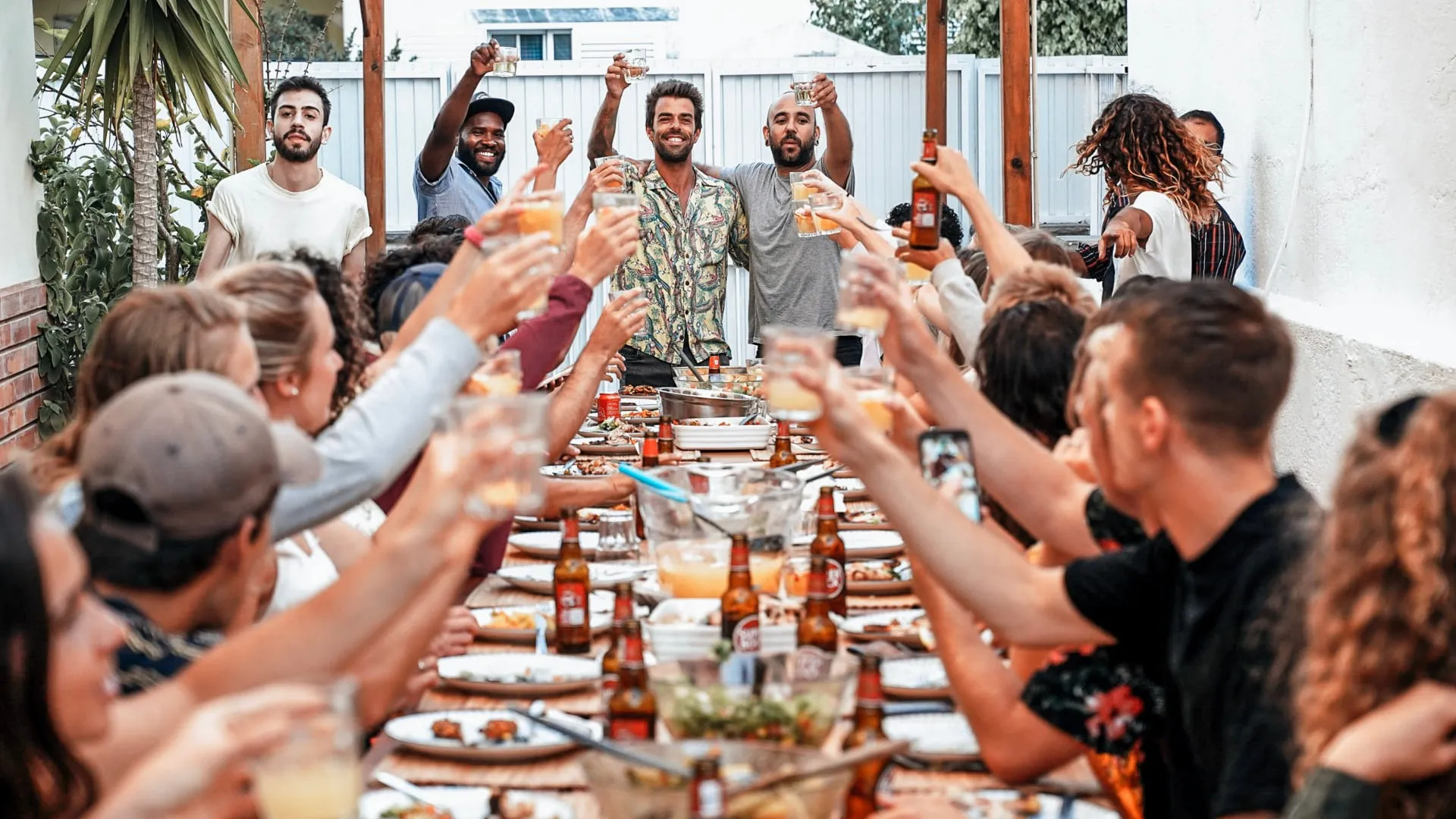 Group of people seated at a long outdoor table raising drinks for a toast during a festive gathering.