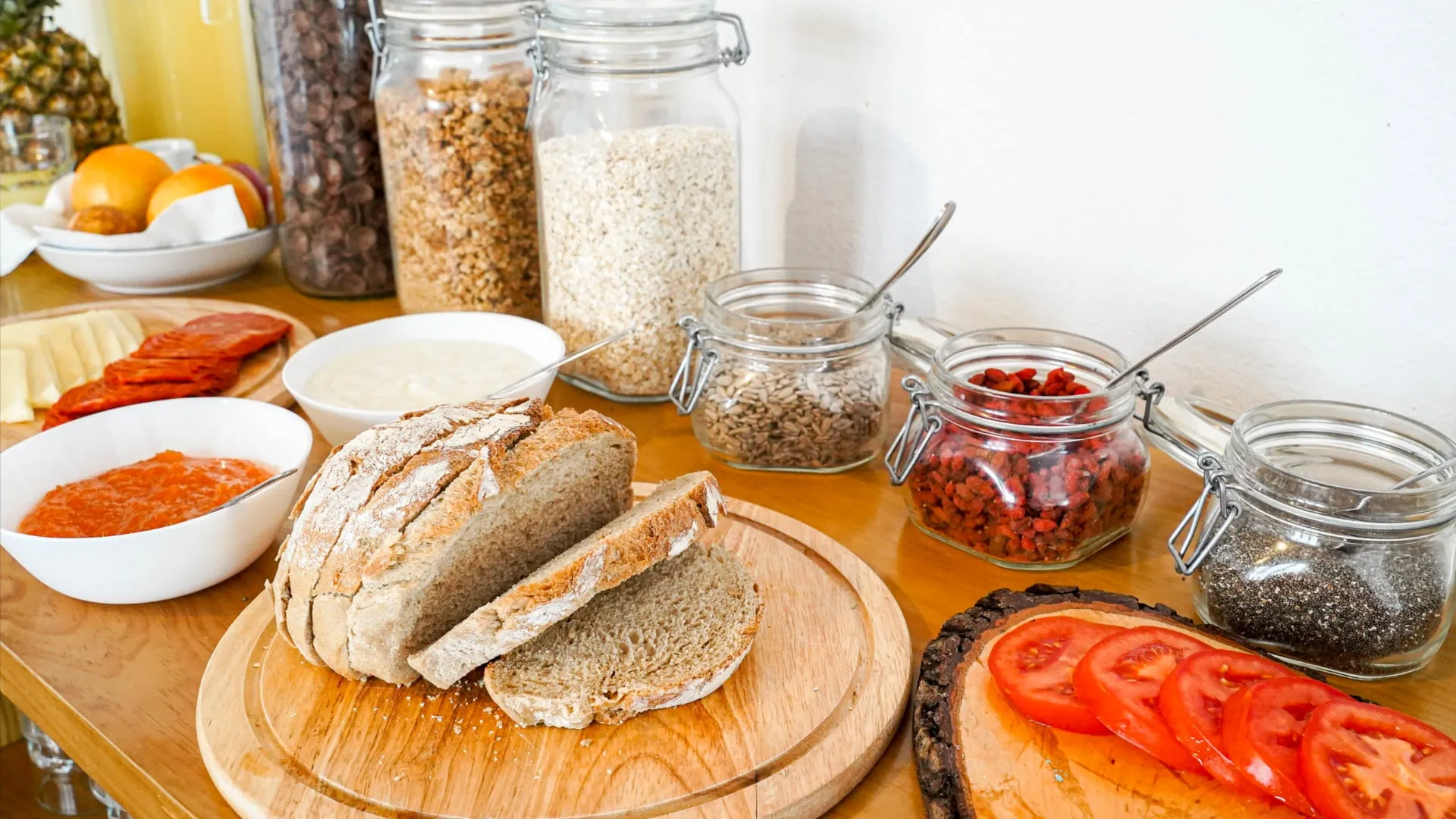 Sliced rustic bread on a wooden board surrounded by jars of seeds, granola, and bowls of spread with tomato slices on bread nearby.