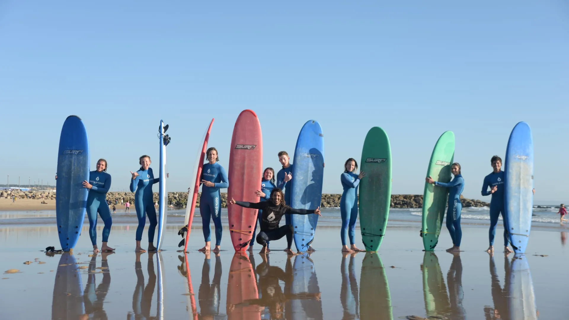 Group of surfers in blue wetsuits holding colorful surfboards standing on a wet sandy beach with reflections.