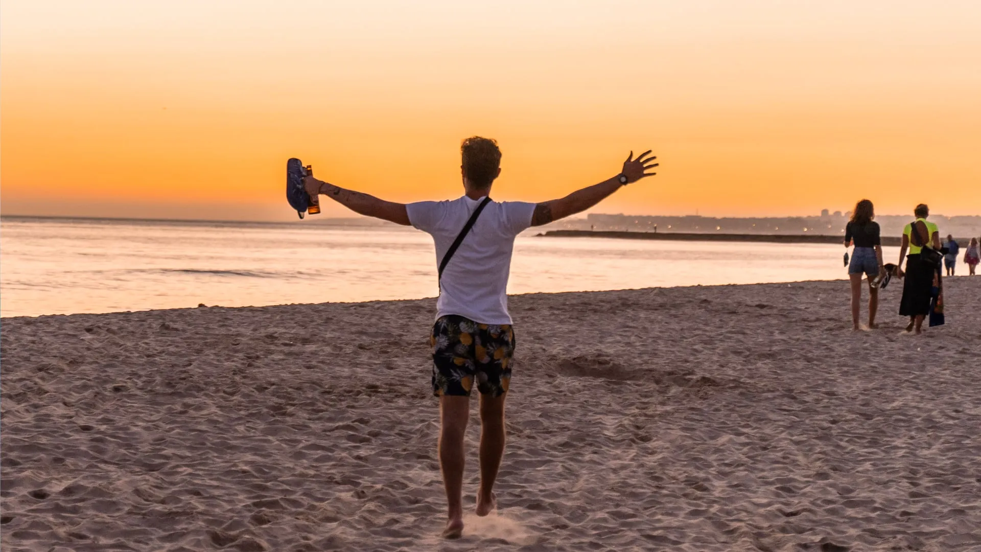 Person with arms outstretched holding shoes and a bottle, walking barefoot on sandy beach at sunset with other people in the distance.