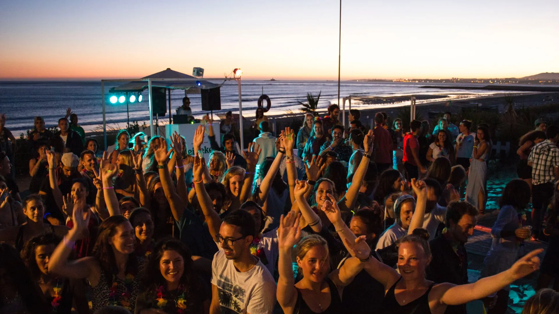 Crowd of people dancing and raising hands at a beach party during sunset with the ocean and coastline in the background.