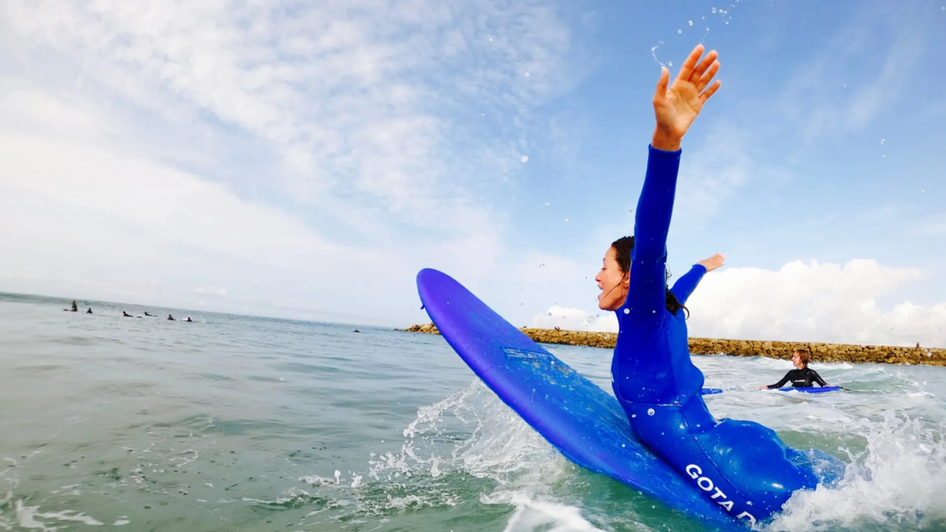 Woman in blue wetsuit excitedly rides a blue surfboard on the ocean near a rocky breakwater.
