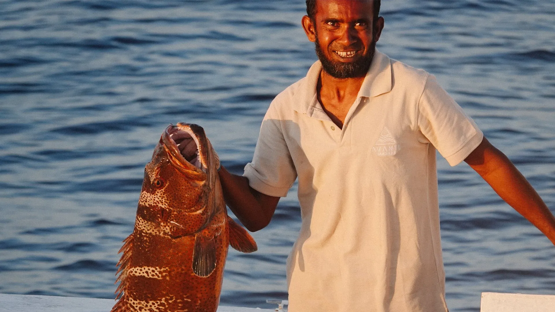 Smiling man in a beige shirt holding a large fish with an open mouth by the water.