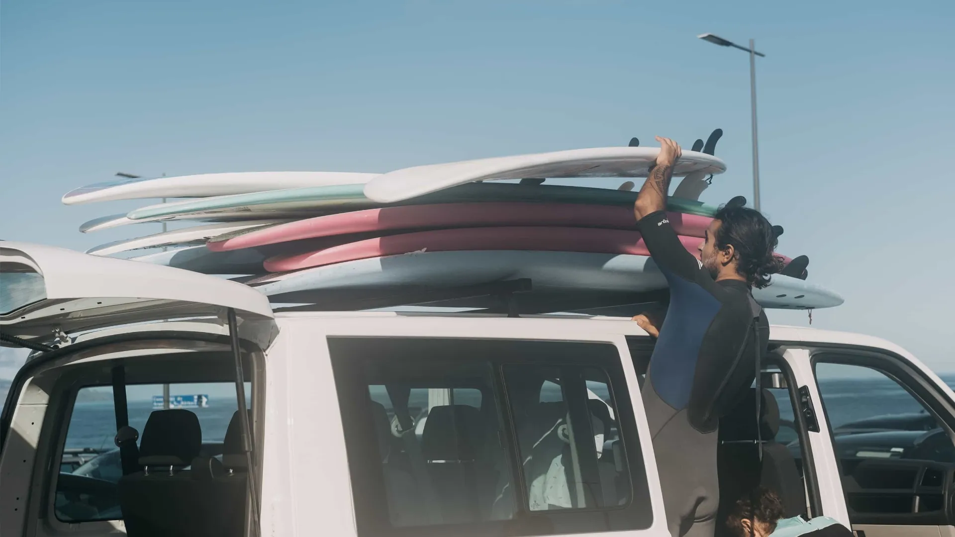 Man in wetsuit securing multiple surfboards stacked on top of a white van near the ocean.