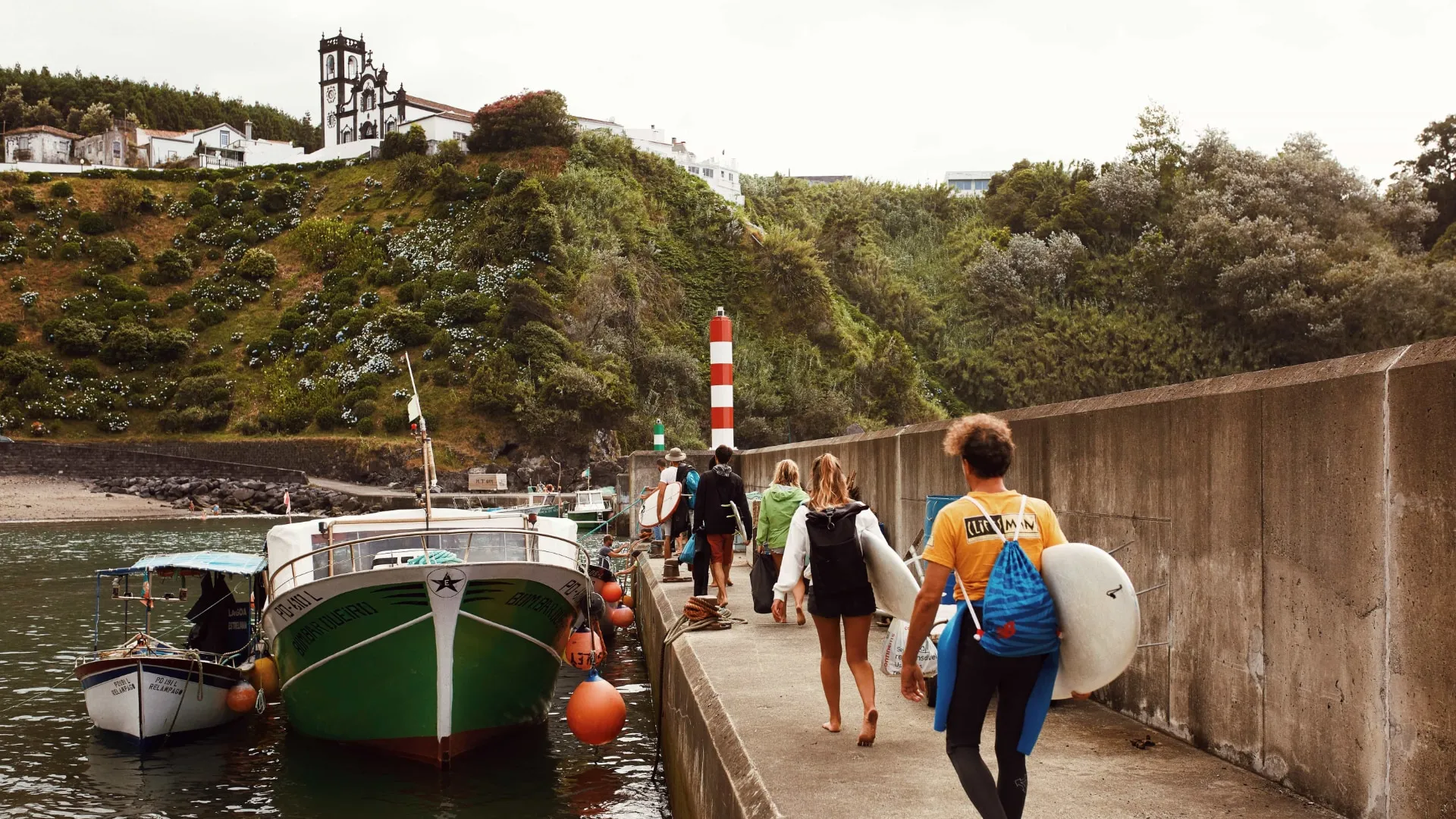 Group of people carrying surfboards walking on a concrete pier beside moored boats with green hillside and buildings in the background.