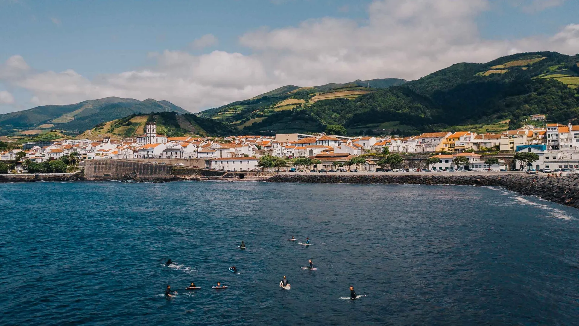 Surfers on boards in the ocean near a coastal town with white buildings and hills in the background under a partly cloudy sky.