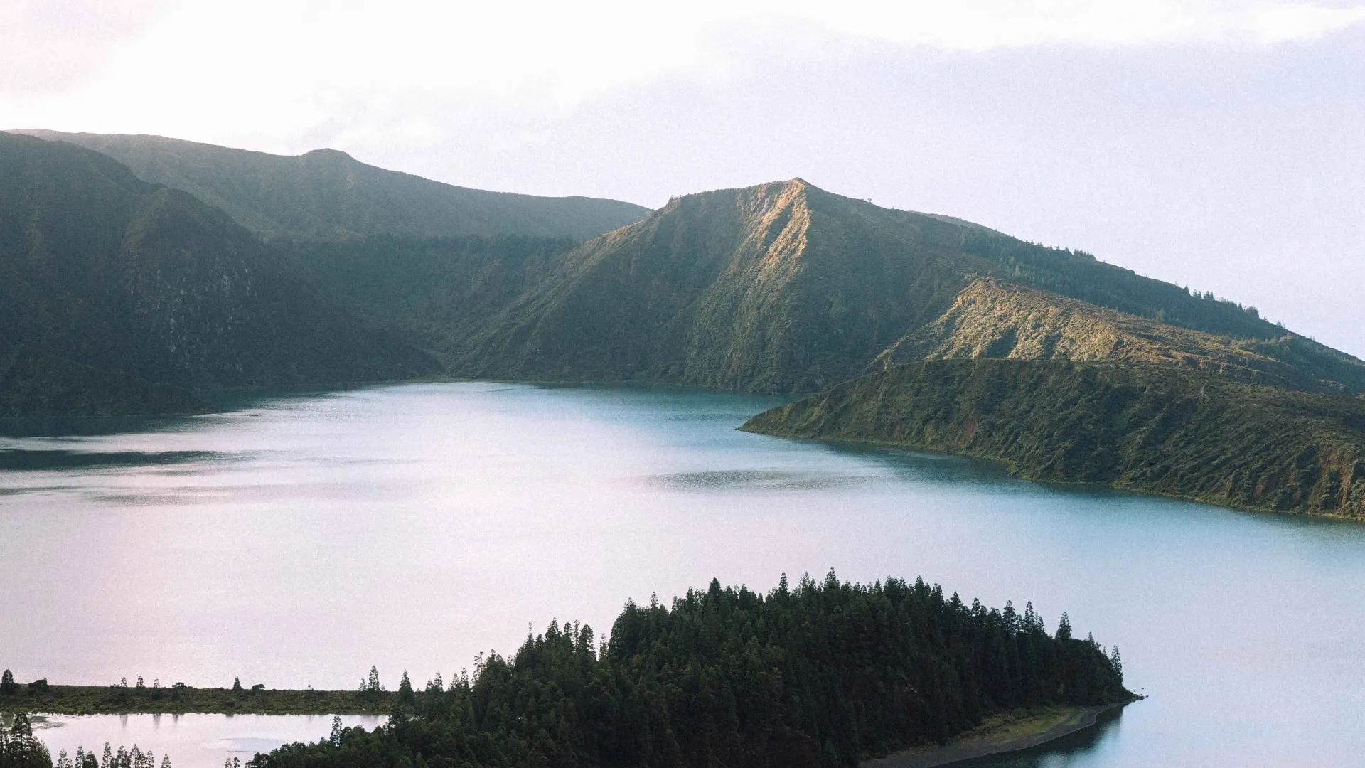 Serene lake surrounded by forested hills and mountains under a bright sky.