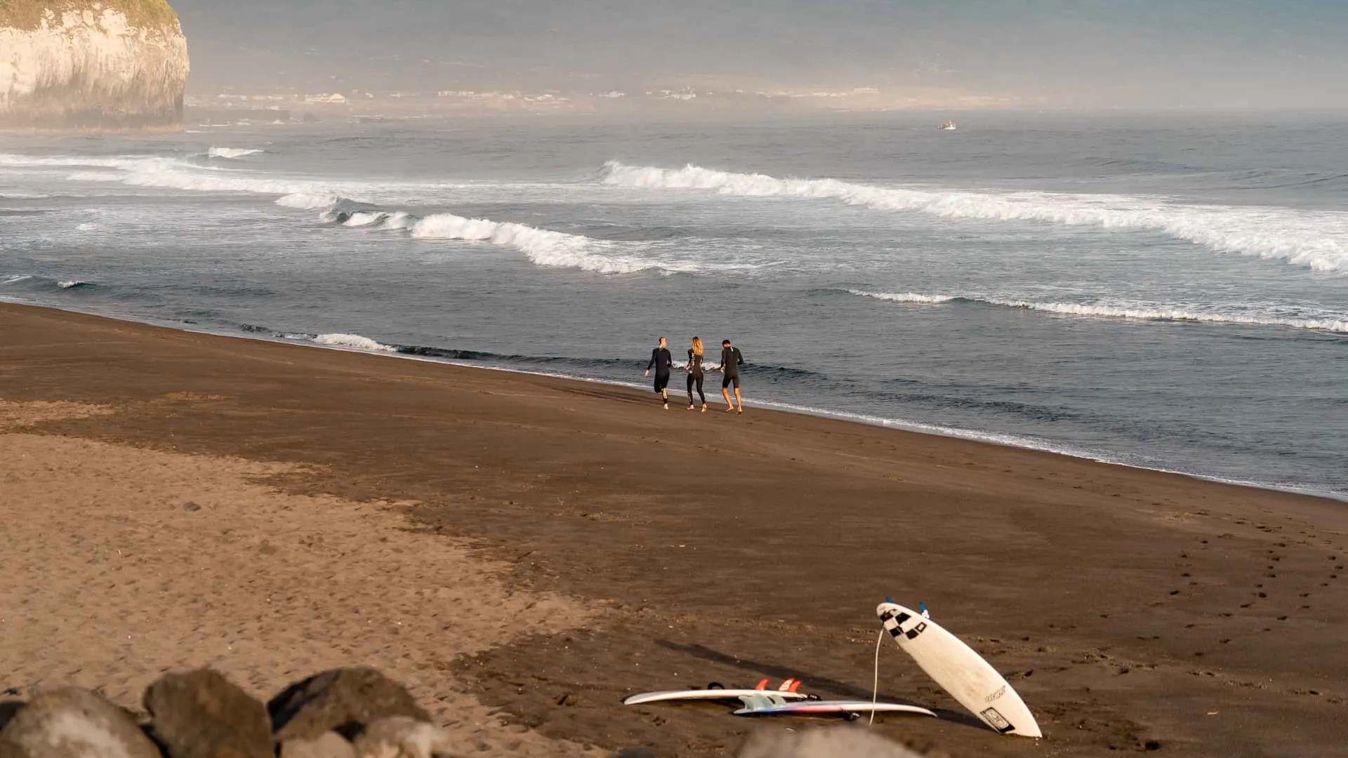 Three people in wetsuits walking along a sandy beach with surfboards lying on the sand and waves in the background.