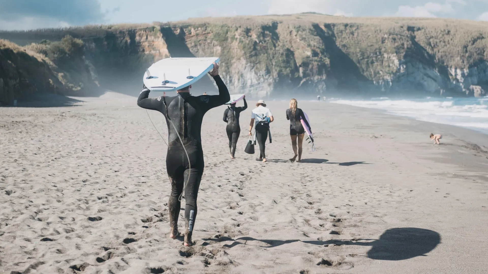 Group of four surfers carrying surfboards walking barefoot on sandy beach towards ocean cliffs.