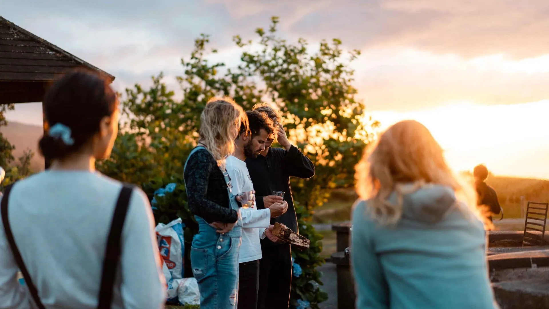 Group of young adults standing outdoors near a fire pit at sunset, with trees and hills in the background.