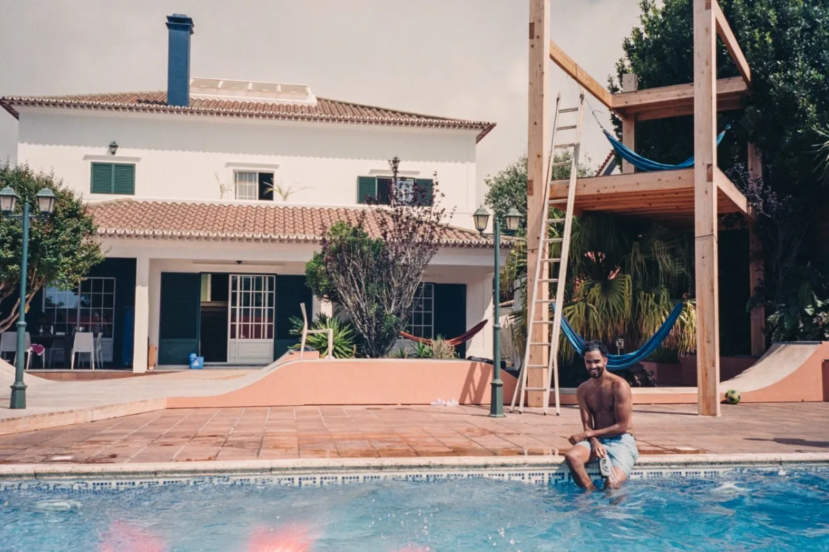 Man sitting at the edge of a swimming pool in front of a white house with a large wooden structure and hammocks.