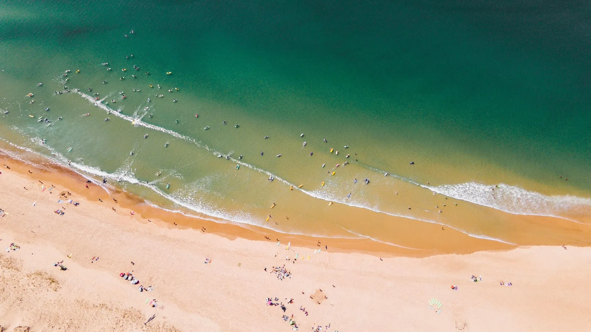 Aerial view of a crowded sandy beach with many people swimming and surfing in the green ocean waves.