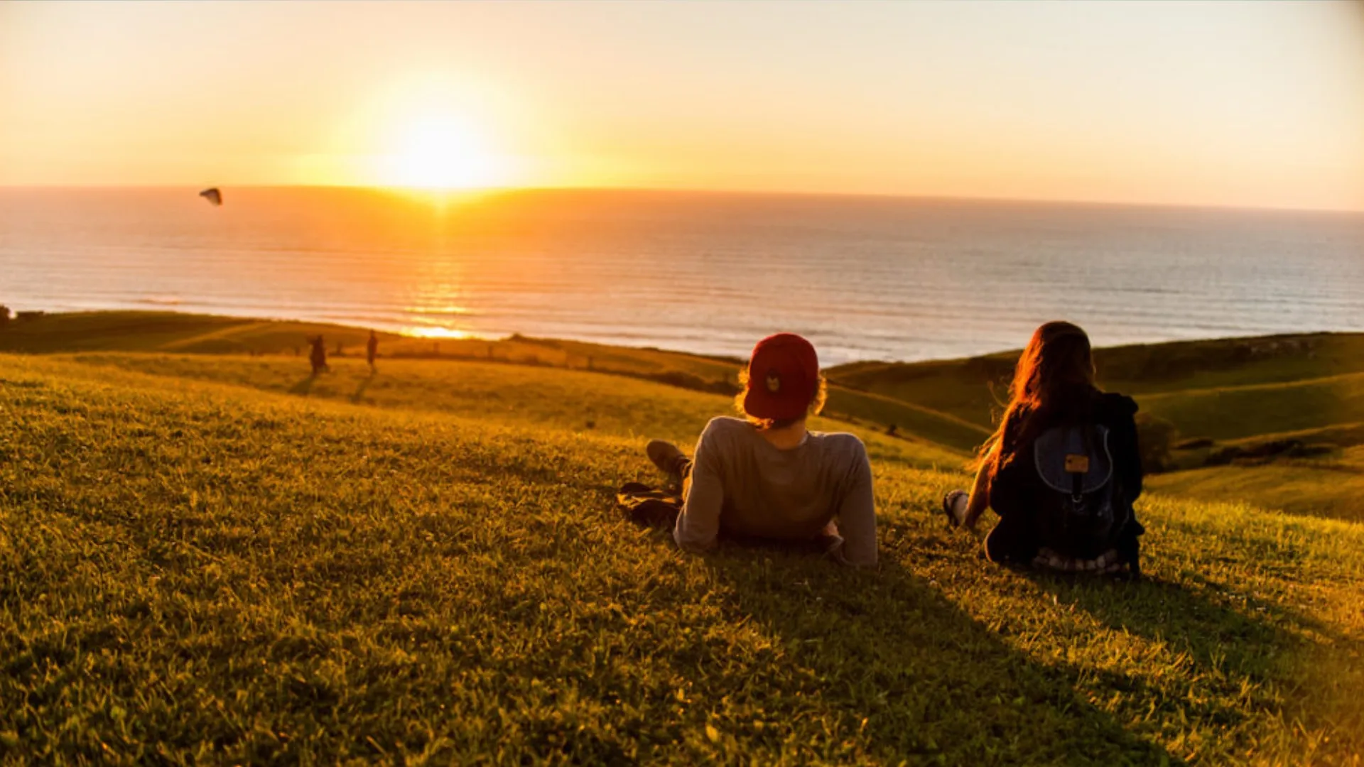 Two people sitting on a grassy hill overlooking the ocean during a golden sunset.