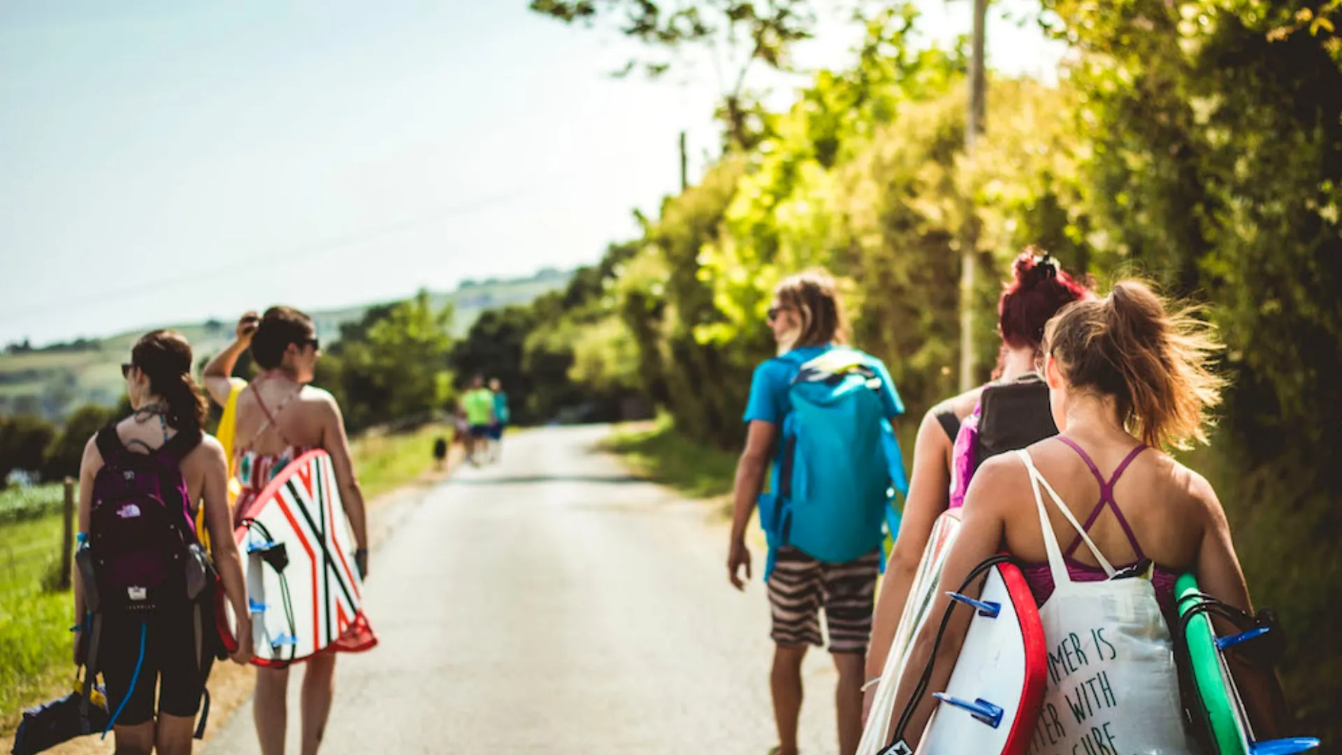Group of young people walking down a sunny road carrying surfboards and backpacks.