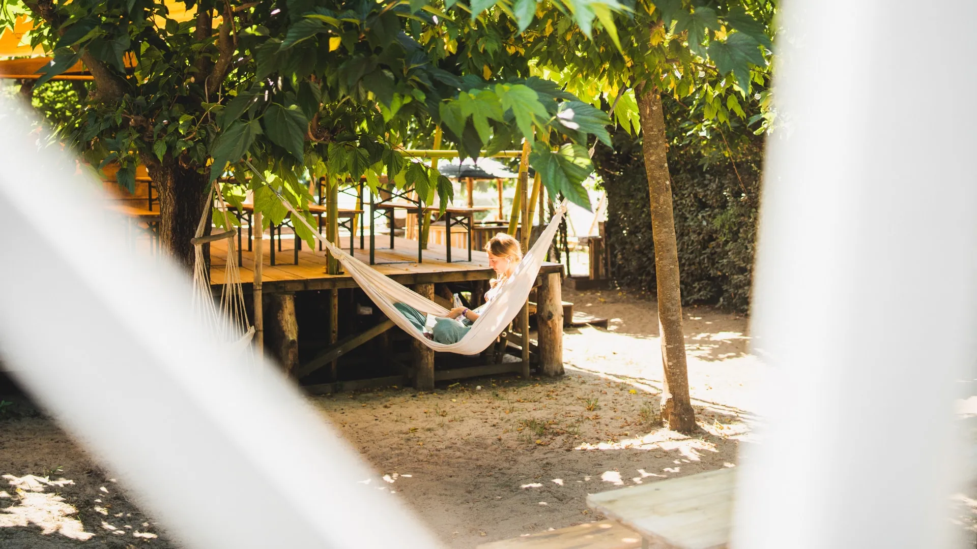 Woman reading a book while sitting in a white hammock under green leafy trees near a wooden deck.