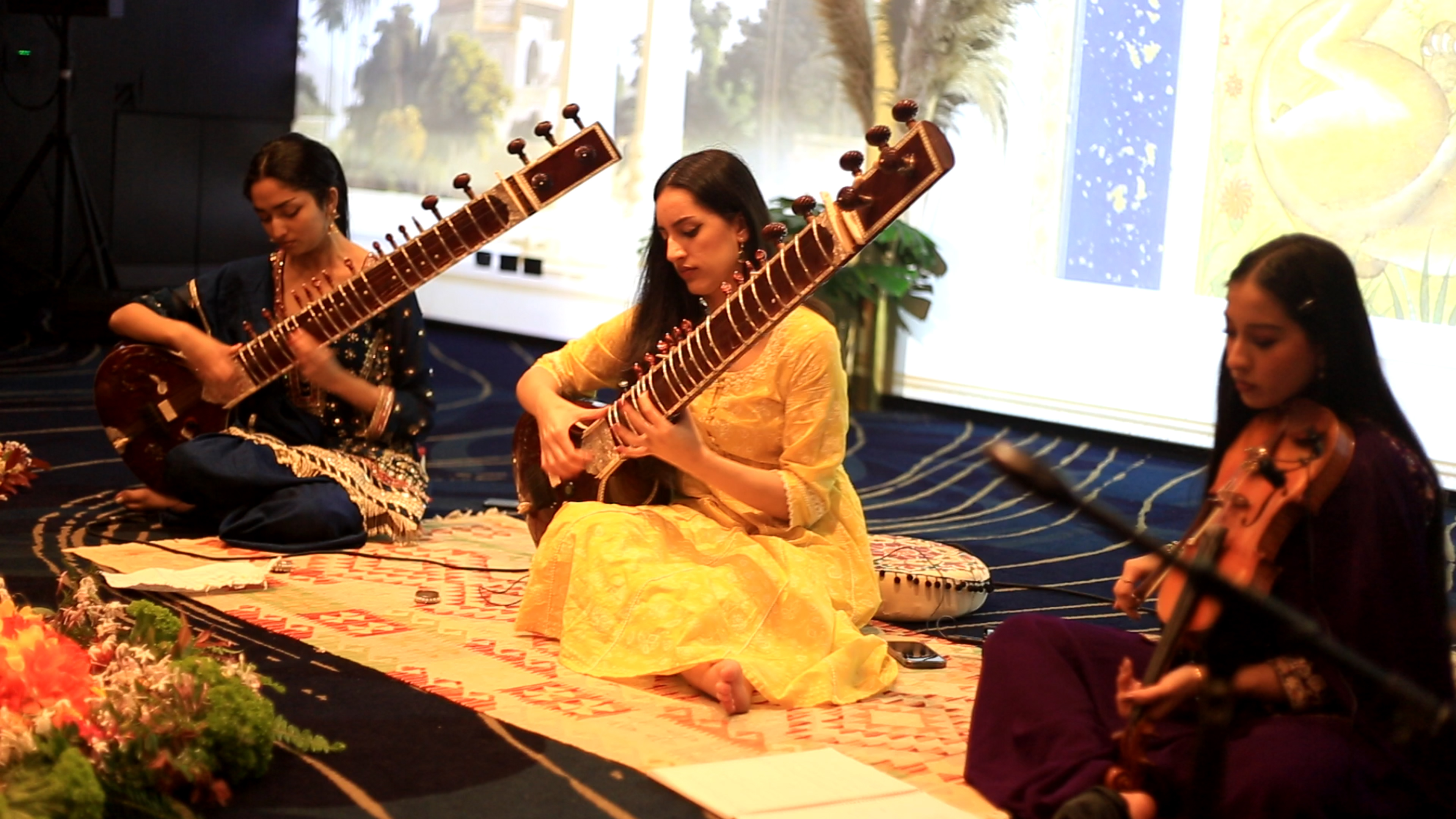 Musicians performing sitar, violin and tabla in front of a projected bird-and-blossom painting