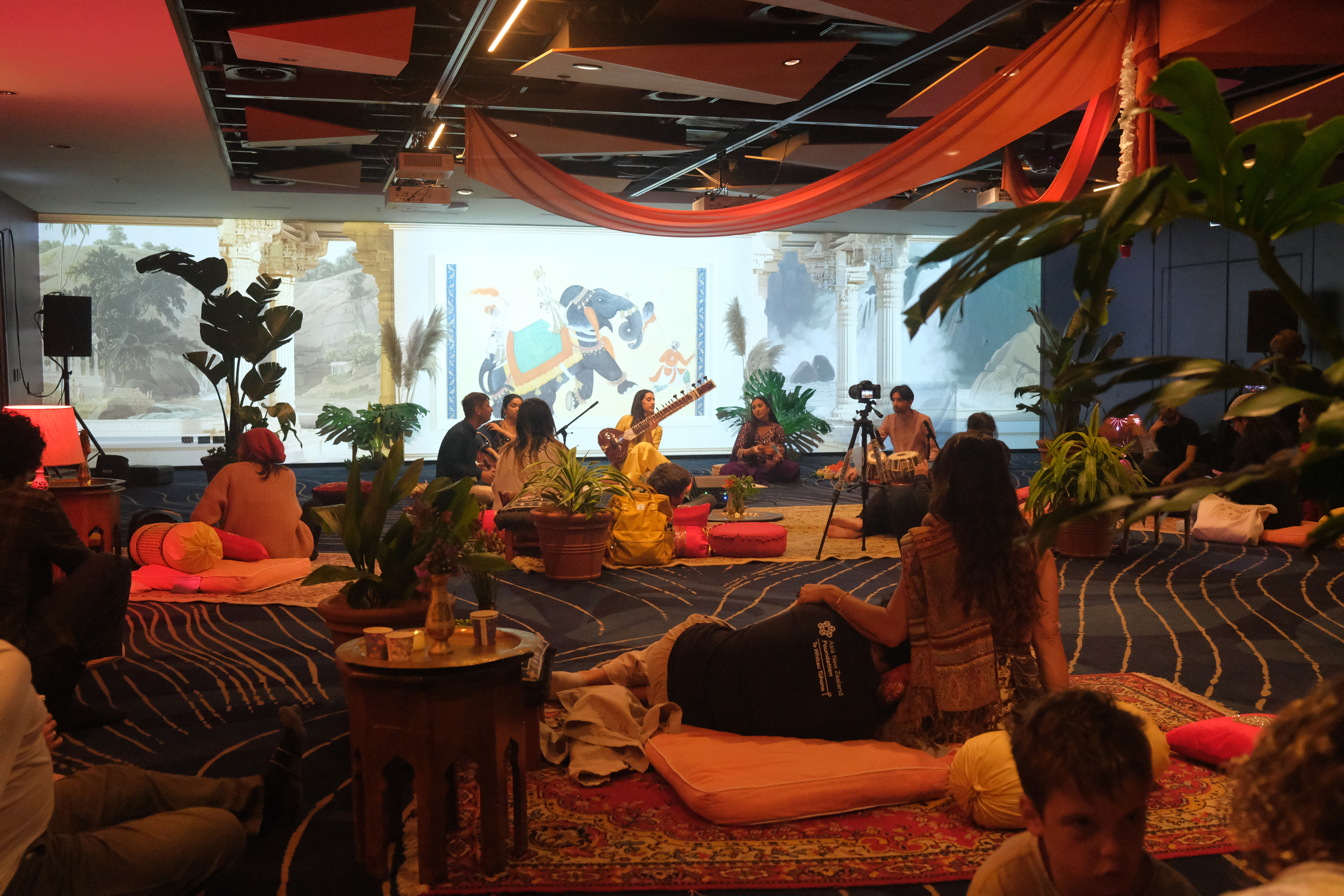 Guests seated on floor cushions as the Basant Ensemble performs beneath draped fabric and a projected Indian miniature painting
