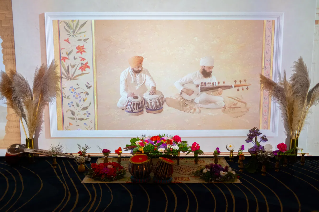 An altar-style display of flowers, tabla drums and a sitar in front of a projected painting of two musicians