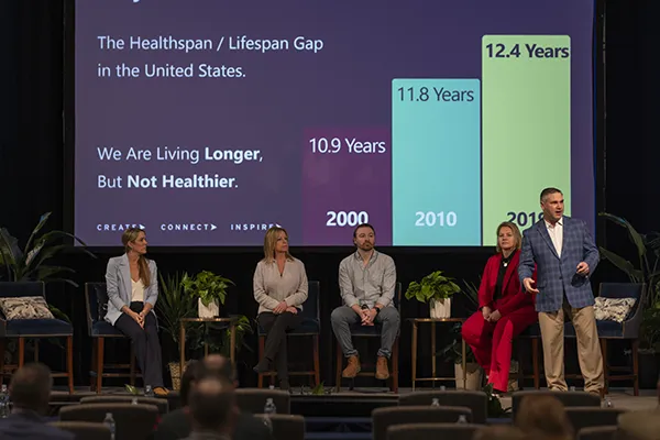 A panel of four seated individuals and one standing presenter on stage during a wellness session at By Design.